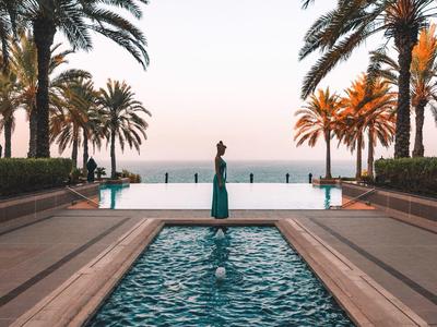 Bazén s vodní fontánou, lemovaný palmami, s výhledem na moře při západu slunce.