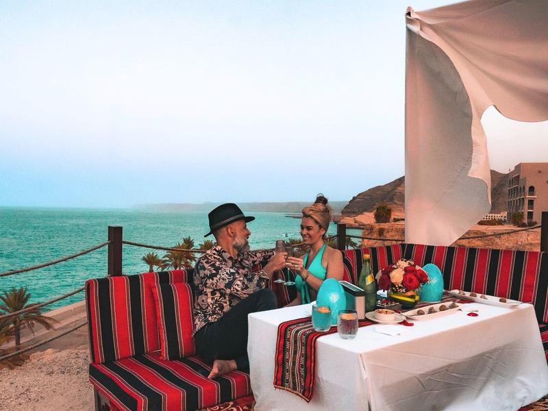 Dva lidé sedí na terase s výhledem na moře, obklopeni barevnými koberci a polštáři.