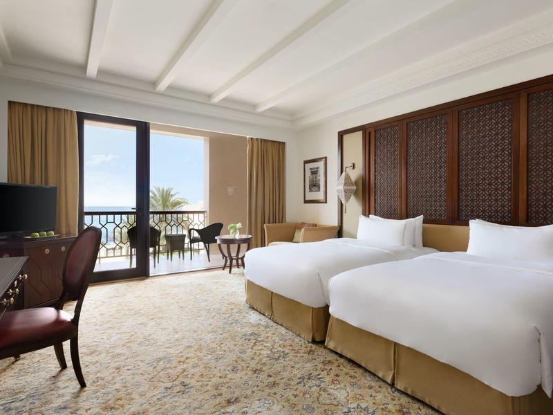 Světly hotelový pokoj se dvěma jednolůžky, balkonem a výhledem na moře.