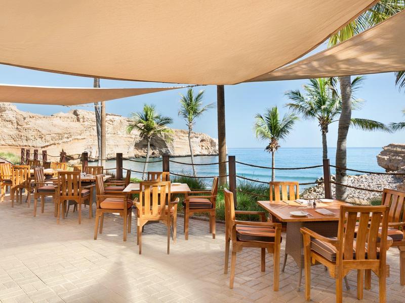 Otevřená terasa s dřevěnými stoly a židlemi, palmami a výhledem na moře pod slunečnou oblohou.