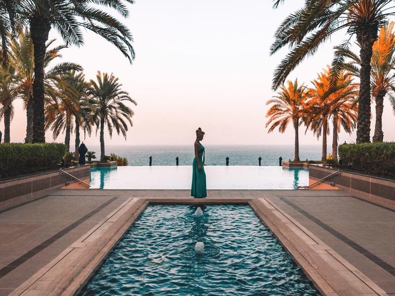 Bazén s vodní fontánou, lemovaný palmami, s výhledem na moře při západu slunce.