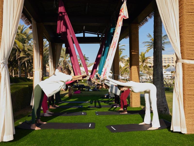 Des personnes pratiquent le yoga aérien dans un espace extérieur entouré de rideaux avec de l'herbe verte.
