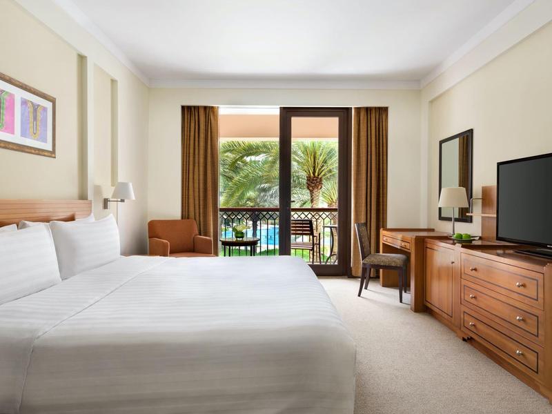 Chambre d'hôtel lumineuse avec lit double, télévision, bureau et balcon avec vue sur des palmiers.