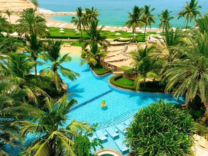 Vue sur une piscine d'hôtel tropicale avec des palmiers et la mer en arrière-plan.