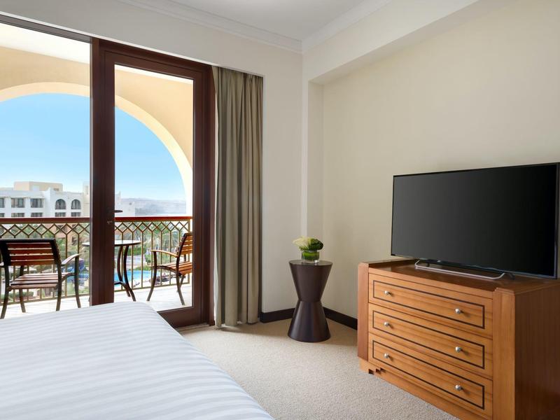 Chambre d'hôtel avec lit, téléviseur sur commode en bois et balcon avec chaises vue sur mer.