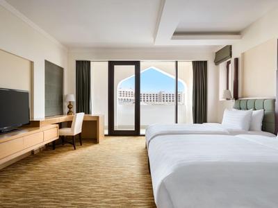 Bright hotel room with two beds, desk, TV, and balcony overlooking the outside view.