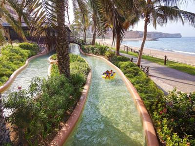 People ride a lazy river beside a beach with palm trees and a promenade.