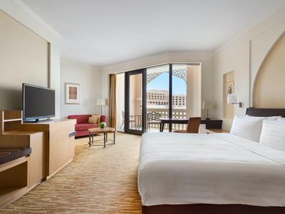 Bright hotel room with large bed, TV, sofa, and balcony overlooking the city.