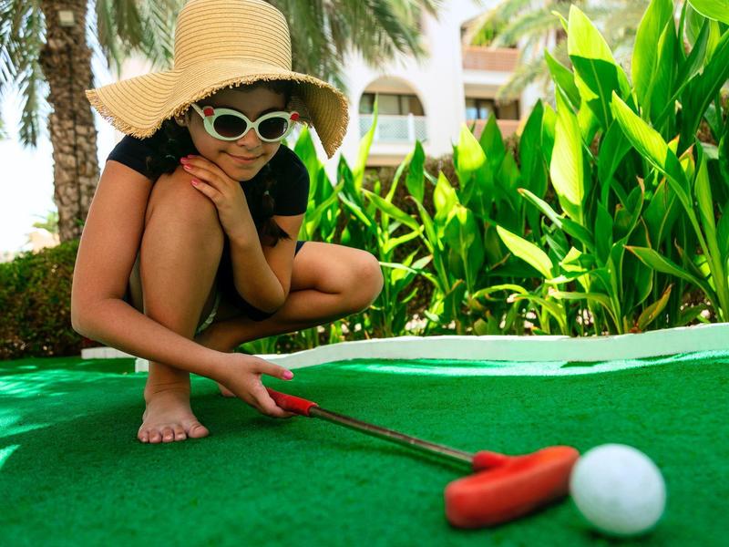Child wearing sunhat and sunglasses playing mini golf outdoors near green plants.