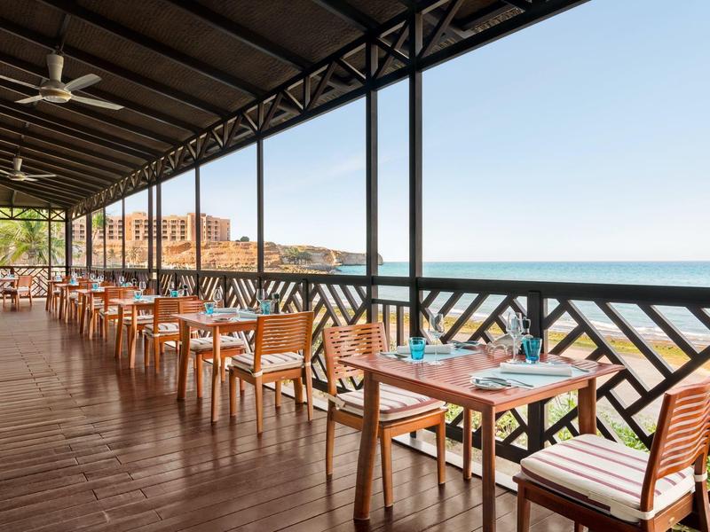 Covered terrace with wooden furniture overlooking the sea and distant buildings.