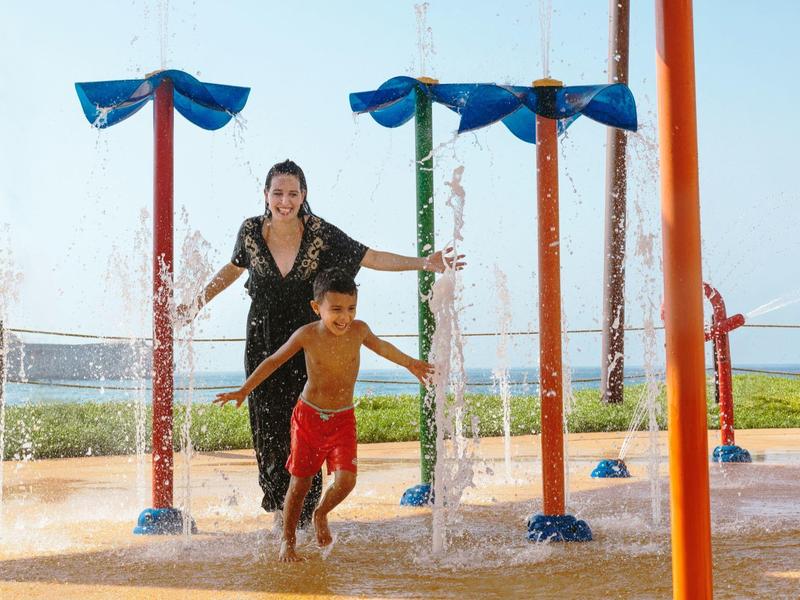 Child and woman laughing and playing under water sprays on an outdoor playground.