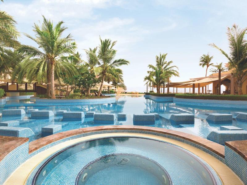 Large pool area with built-in hot tub surrounded by palm trees under a blue sky.