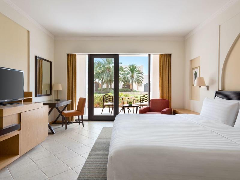 Bright hotel room with double bed, desk, TV, and balcony overlooking palm trees.