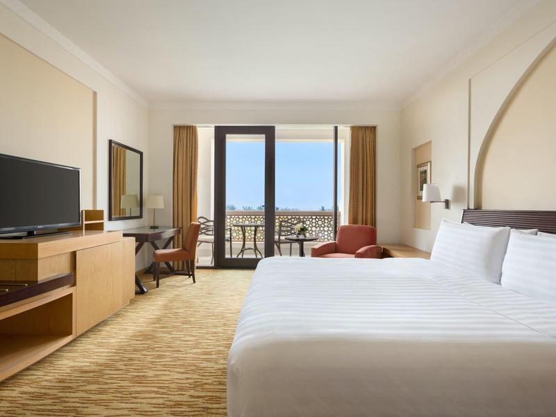 Bright hotel room with large bed, TV, desk, and balcony overlooking the city.