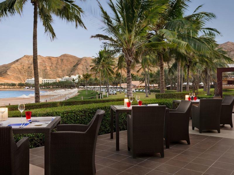 Hotel terrace with tables, wicker chairs, palm trees, and mountain view by the beach.