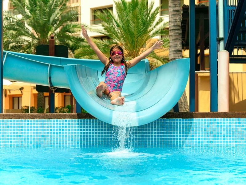 A girl joyfully slides down a water slide into a pool.