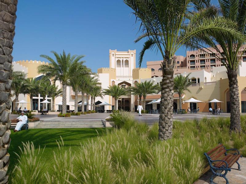 Hotel grounds with palm trees, lawns, and benches under clear blue sky.