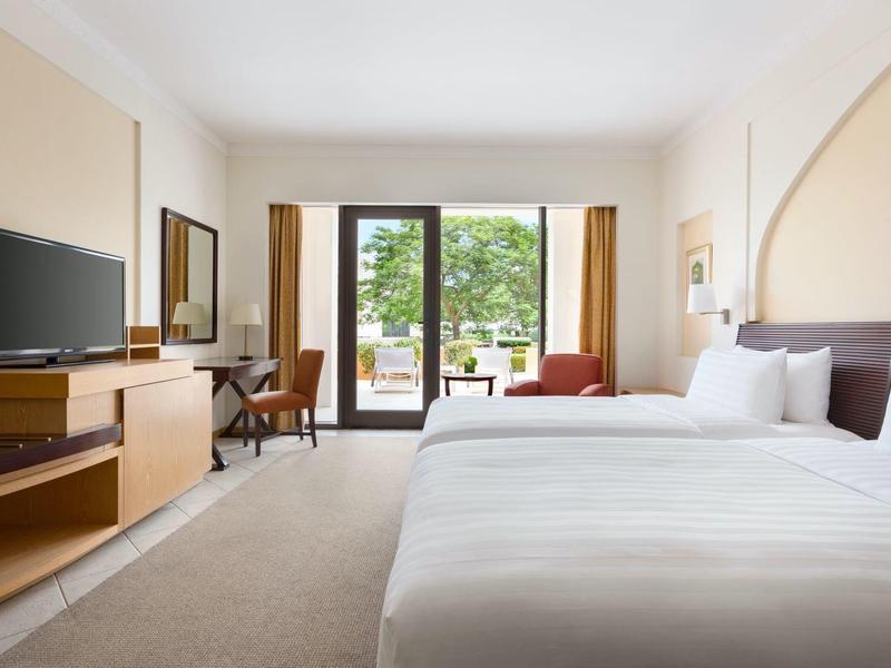 Bright hotel room with large bed, TV, desk, and balcony overlooking trees.