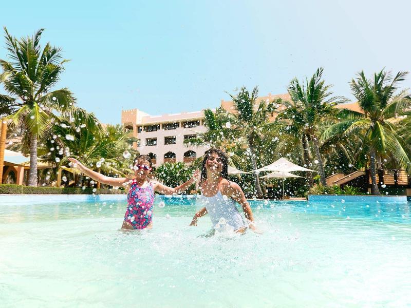 Two children playing in the shallow pool in front of a hotel with palm trees in the background.