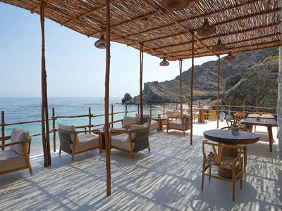 Offene Terrasse mit Holzmöbeln und Blick auf das Meer und die Felsen im Hintergrund.