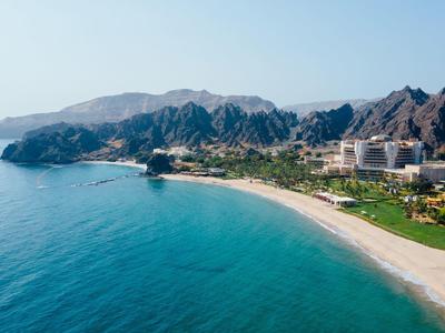 Küste mit blauem Meer, weißem Sandstrand und grünen Flächen vor bergigem Gelände unter blauem Himmel.