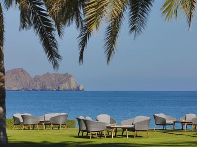 Césped verde con grupos de asientos bajo palmeras con vista al mar azul y rocas distantes.