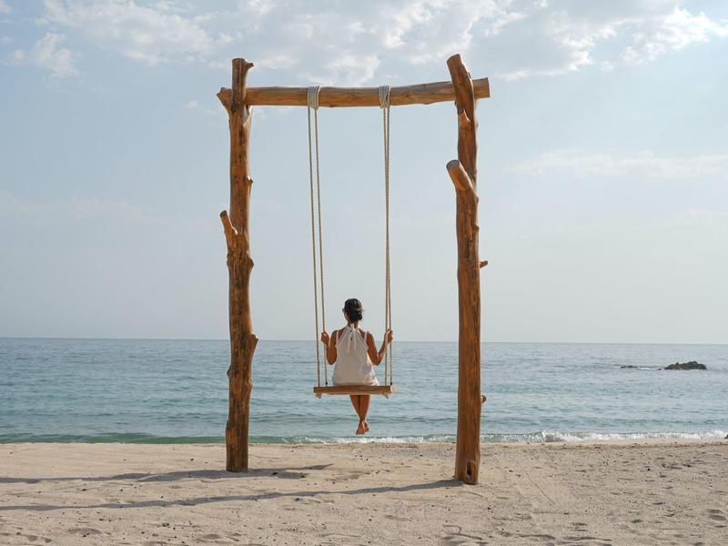 Persona sentada en un columpio de madera en la playa con vista al mar tranquilo.