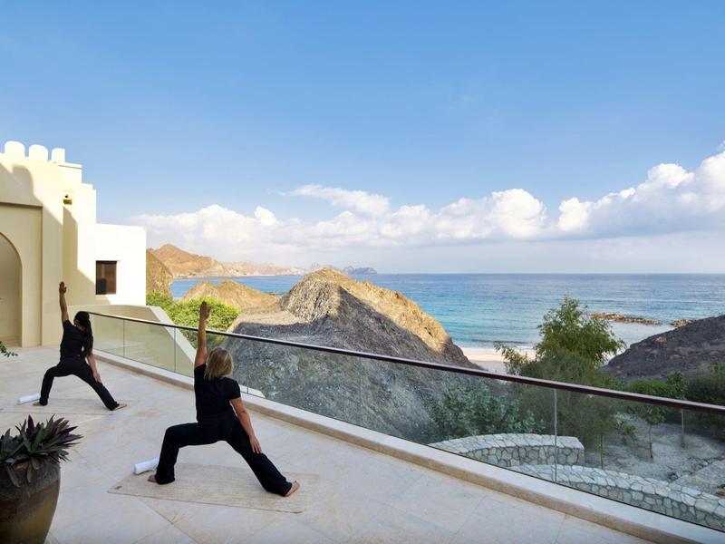 Dos personas hacen yoga en una terraza con vista al mar y rocas de fondo.