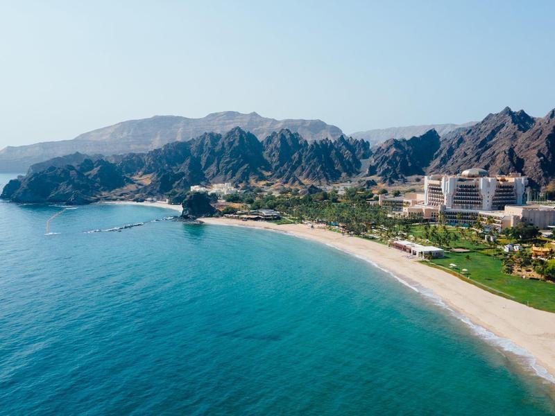 Costa con playa de arena, agua azul y montañas rocosas, hoteles y vegetación verde.