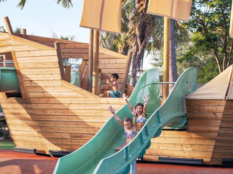 Niños descienden por un tobogán verde en una piscina con un barco de madera de fondo.