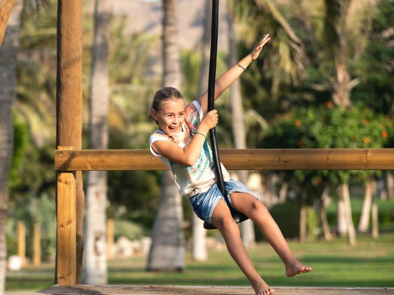 Niña jugando en un columpio de cuerda en un parque con palmeras y montañas al fondo.