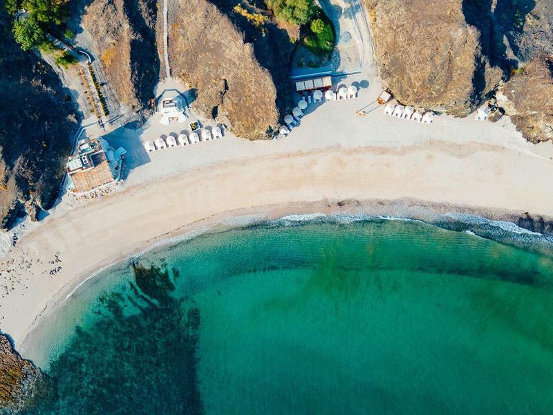 Vista aérea de una cala costera con agua verde clara y playa arenosa entre rocas.