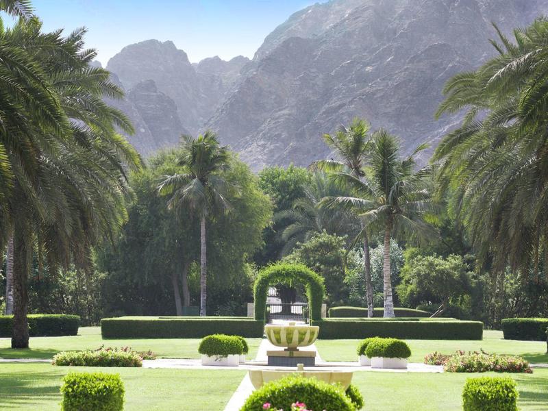 Gran jardín con palmeras, fuente y montañas al fondo en un complejo hotelero.