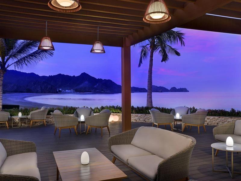 Terraza de hotel al atardecer con sofás, mesas y palmeras con vistas al mar y montañas.