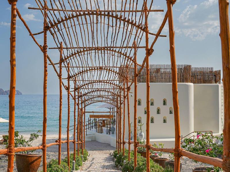 Un camino arqueado de metal oxidado conduce a un edificio blanco con cielo y mar al fondo.