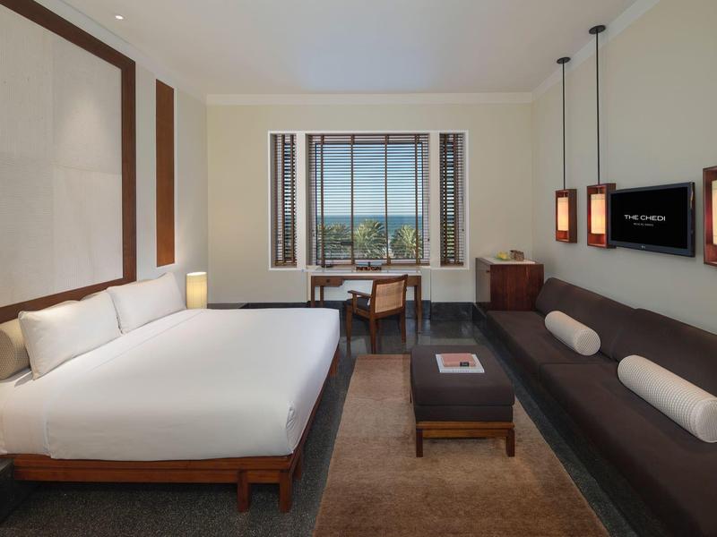 Moderní hotelový pokoj s velkou postelí, pohovkou, psacím stolem a výhledem na zeleň.