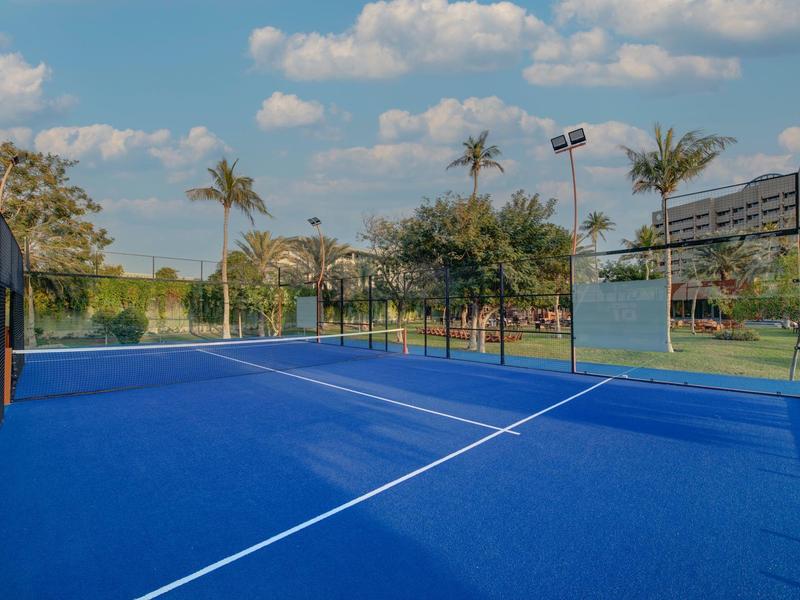 Court de tennis extérieur bleu avec des palmiers et un ciel nuageux en arrière-plan.