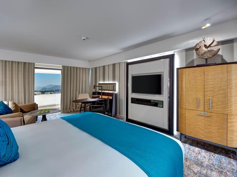 Chambre d'hôtel lumineuse avec grand lit, chemin de lit bleu, canapé et grande fenêtre avec vue sur le lac.