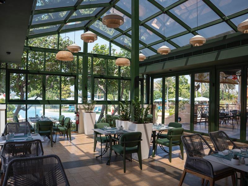 Véranda lumineuse et ouverte avec tables, chaises et vue sur piscine extérieure et jardin.