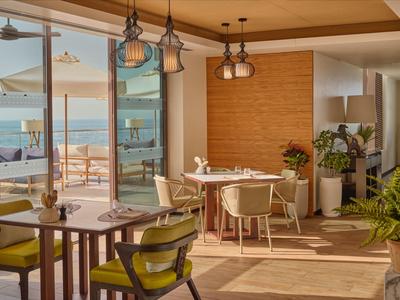 Espace restaurant lumineux avec meubles en bois et vue sur la mer à travers de grandes fenêtres.