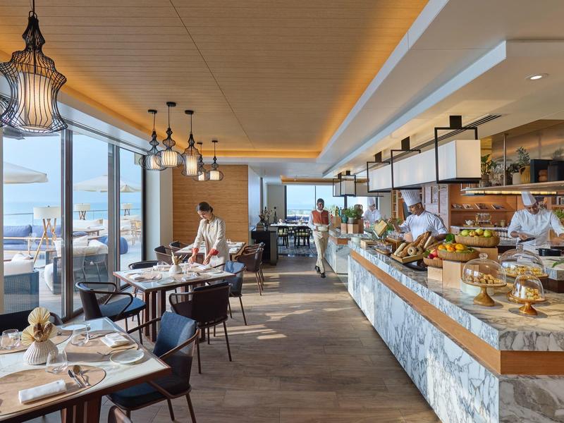 Restaurant moderne avec grandes fenêtres, mobilier élégant et long buffet avec des chefs.