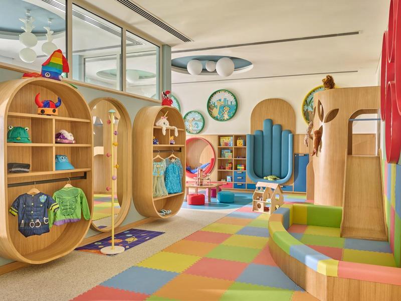 Salle de jeux colorée pour enfants avec étagères en bois, tapis multicolore et coin salon lumineux.