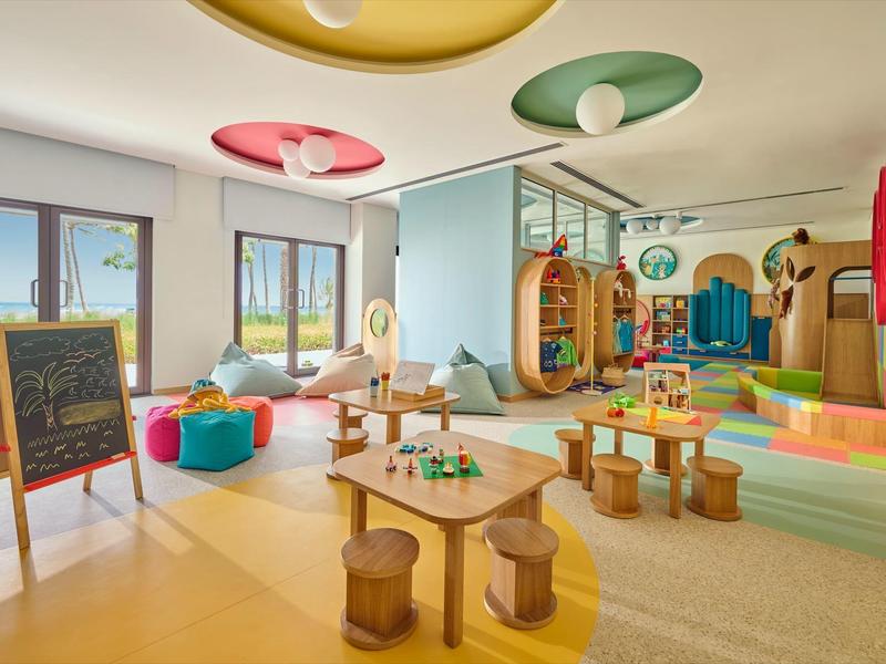 Salle de jeux colorée pour enfants avec tables, poufs, tableau noir et formes douces dans un espace lumineux.