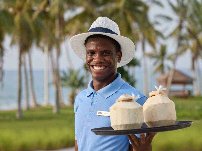 Homme souriant portant un chapeau servant des noix de coco sur un plateau dans un cadre tropical.