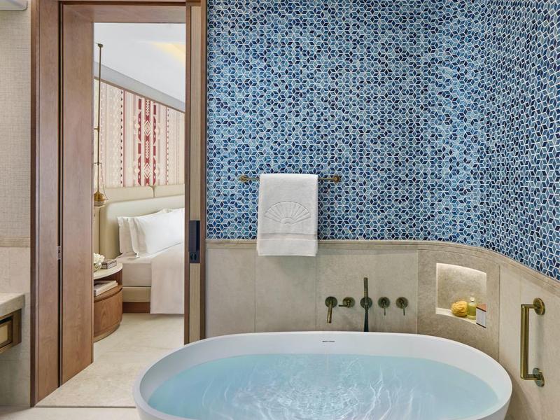 Baignoire moderne remplie d'eau devant un mur en mosaïque grise dans une salle de bain lumineuse à côté d'un cadre de porte en bois.