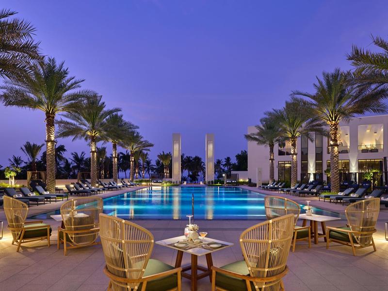 Espace piscine élégant d'un hôtel avec palmiers et sièges confortables au crépuscule.