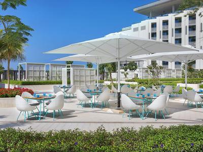 Espace extérieur moderne avec chaises blanches, tables bleues et grands parasols devant un hôtel.