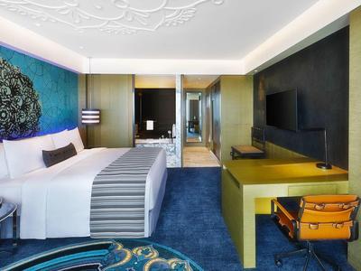 Moderní hotelový pokoj s modrým kobercem, manželskou postelí a psacím stolem s oranžovou židlí.