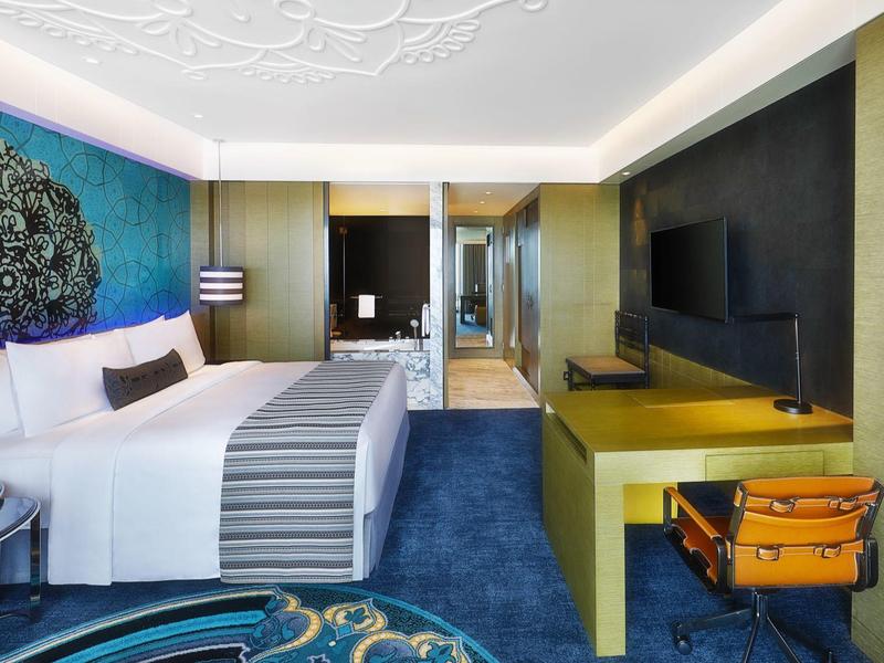 Moderní hotelový pokoj s modrým kobercem, manželskou postelí a psacím stolem s oranžovou židlí.