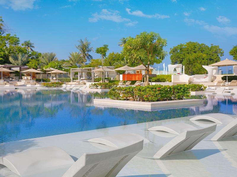 Velký bazén s bílými lehátky a slunečníky v tropickém hotelu pod modrou oblohou.